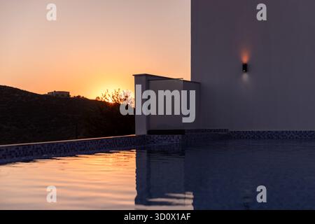 Un coucher de soleil serein baigne une piscine sur le toit d'une lumière chaude, reflétant des couleurs douces sur l'eau plate. Les murs de villa blancs modernes encadrent la scène, créant une paix Banque D'Images