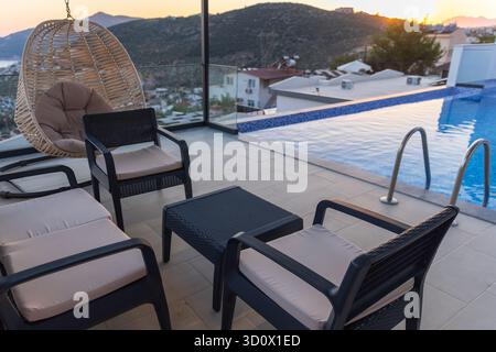 Un salon reposant sur le toit en bordure de piscine avec des sièges rembourrés, une chaise suspendue et une balustrade en verre offre une vue sereine sur le coucher du soleil sur la ville et les collines, idéal Banque D'Images