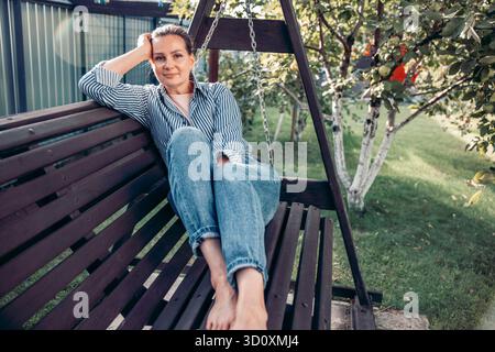 Une jeune femme caucasienne aux cheveux bruns se détend sur une balançoire dans un jardin. Elle porte des vêtements décontractés en denim et profite d'un moment paisible entouré de gre Banque D'Images