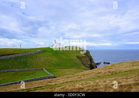 Un phare blanc se trouve au sommet de falaises herbeuses surplombant une mer calme. Des murs de pierre sinueux et un chemin incurvé encadrent la scène, tandis que des nuages d'humeur changeante ajoutent un tim Banque D'Images