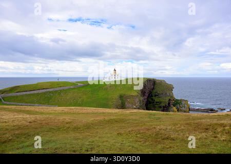 Un phare blanc se trouve au sommet de falaises herbeuses surplombant une mer calme. Des murs de pierre sinueux et un chemin incurvé encadrent la scène, tandis que des nuages d'humeur changeante ajoutent un tim Banque D'Images