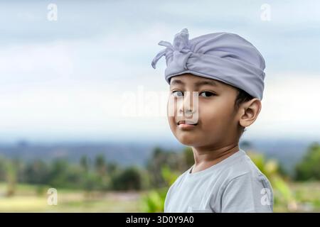 Portrait d'un enfant balinais en bandeau cérémonial blanc "Udeng" et chemise, debout à l'extérieur avec un fond de champ de riz flou le jour du Galungan. Banque D'Images