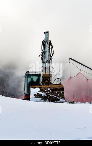 Un chantier de construction enneigé avec une excavatrice. La machine est positionnée sur une pente, entourée d'une atmosphère brumeuse et d'un filet de sécurité rouge. Banque D'Images