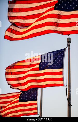 Drapeaux américains flottant à la lumière dorée de l’heure d’or à Washington DC | WASHINGTON D.C., États-Unis — Des drapeaux américains qui se chevauchent remplissent le cadre à la lumière dorée de l’heure d’or près du Washington Monument, leurs étoiles et leurs rayures brillant contre un ciel doux du soir. Les drapeaux font partie d’un anneau de cinquante drapeaux autour de la base du monument, chacun représentant un État américain, entretenu toute l’année par le National Park Service. La scène reflète la lumière de fin de journée commune sur le National Mall dans les minutes précédant le coucher du soleil. Banque D'Images