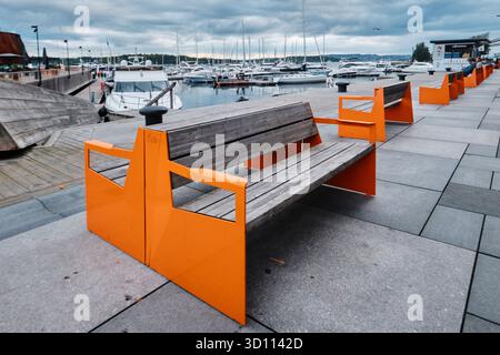 Oslo, Norvège - 8 septembre 2025 : bancs vides en bois de couleur orange sur le remblai et bateaux amarrés dans le port du district d'Aker Brygge Banque D'Images