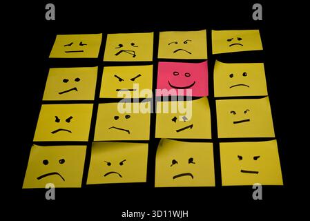 15 papiers jaunes placés parallèlement les uns aux autres avec des visages dessinés sur eux exprimant la colère ou la tristesse, face à un papier rose avec un visage exprimant le happin Banque D'Images