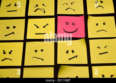 15 papiers jaunes placés parallèlement les uns aux autres avec des visages dessinés sur eux exprimant la colère ou la tristesse, face à un papier rose avec un visage exprimant le happin Banque D'Images