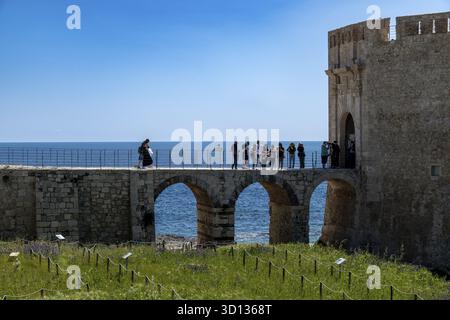 Syrakus, Sizilien - Italie - 04-09-2025 : groupe de touristes marchant et debout sur un pont de pierre historique avec une base voûtée, relié à une côte pour Banque D'Images