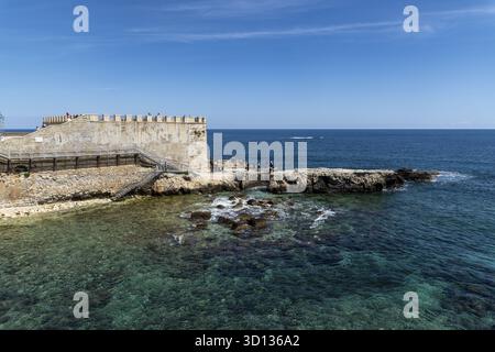 Syrakus, Sizilien - Italie - 04-09-2025 : une plate-forme en pierre altérée pénètre dans la mer turquoise de Syrakus, avec des visiteurs debout près du bord en dessous Banque D'Images