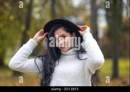 Belle jeune femme brune dans un pull blanc et chapeau noir dans le parc d'automne Banque D'Images