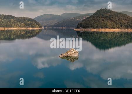 Eau sereine du lac reflétant les montagnes environnantes et la forêt luxuriante, avec une croix encastrée dans une formation rocheuse solitaire Banque D'Images
