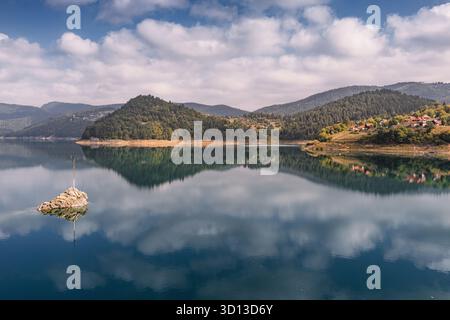 Eau sereine du lac reflétant les montagnes environnantes et la forêt luxuriante, avec une croix encastrée dans une formation rocheuse solitaire Banque D'Images