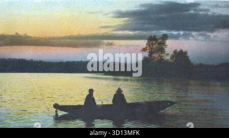 Deux personnages sont assis tranquillement dans une barque alors que le crépuscule s'installe sur un lac calme. Le ciel brille avec une lumière qui s'estompe — orange, jaune et bleu — tandis que les nuages sombres dérivent au-dessus. Les arbres bordent le rivage lointain en silhouette. Intitulée Evening Shadows, cette carte postale 1908 de Russell Brothers capture un moment de calme, de réflexion et de beauté naturelle. D'une carte postale originale. Banque D'Images
