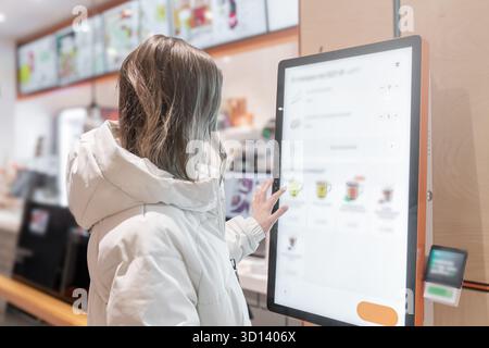 Une femme utilise un kiosque libre-service dans un café moderne, mettant en évidence la commodité technologique dans le dîner, Une femme en veste blanche choisit des boissons et commande de la nourriture Banque D'Images