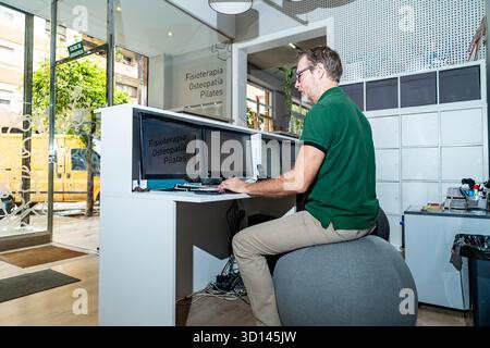 Homme assis sur un ballon d'équilibre, tapant sur ordinateur dans un bureau de clinique de physiothérapie Banque D'Images