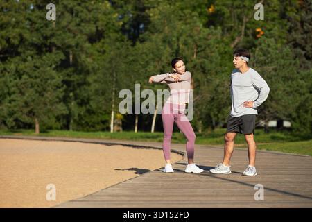 Jeune femme asiatique étirant les bras tout en se tenant sur un chemin en bois à l'extérieur à côté d'un homme caucasien d'âge moyen avec les mains sur les hanches, les deux portant des chaussures de sport, des arbres en arrière-plan Banque D'Images