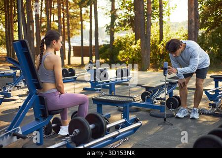 Jeune femme caucasienne assise sur le banc d'entraînement se préparant à l'exercice tandis que jeune homme caucasien, ajustant le smartphone sur trépied dans la salle de gym extérieure entourée d'un équipement d'haltérophilie Banque D'Images