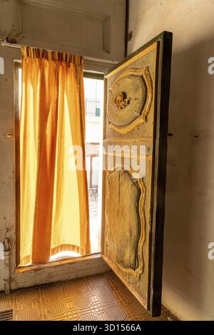 Scicli, Sizilien - Italie - 04-05-2025 : une vieille porte en bois sculpté de l'église San Giovanni partiellement ouverte avec la lumière du soleil coulant à travers une curta orange Banque D'Images
