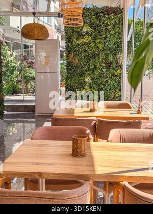 Un intérieur de restaurant confortable avec des tables en bois et des chaises roses douces. Un mur vert de plantes ajoute une touche naturelle au décor moderne. Banque D'Images