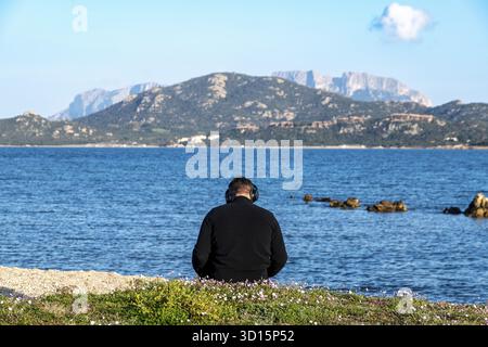 Olbia - Sardinien - Italien - 02-18-2025 : homme avec des écouteurs assis sur une prairie côtière, face à la mer calme près d'Olbia, avec des montagnes escarpées dans le th Banque D'Images