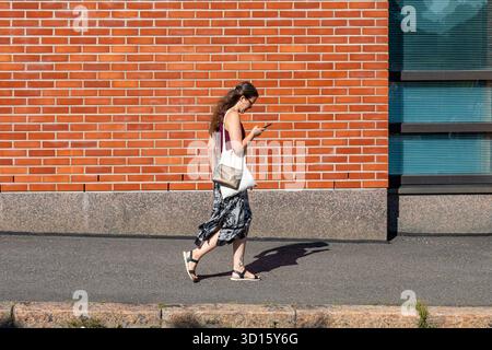Jeune femme naviguant sur le téléphone tout en se promenant le long de ENSI Linja lors d'une soirée d'été ensoleillée et chaude dans le quartier de Kallio à Helsinki, Finlande Banque D'Images