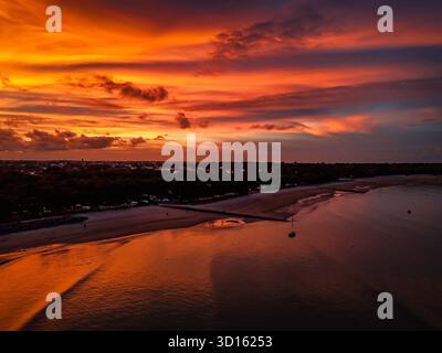 Un coucher de soleil dramatique peint le ciel en orange et rouge tandis que les eaux calmes reflètent les teintes flamboyantes. Des arbres silhouettés bordent la côte tandis qu'une jetée et un har lointain Banque D'Images