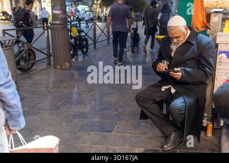 Jérusalem, Israël, 23 octobre 2025 Un homme âgé à la longue barbe, vêtu d'un manteau noir et d'une kippa blanche, est assis sur un tabouret, regardant un petit smartphone dessus Banque D'Images
