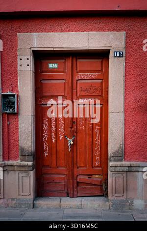 Double porte en bois rouge verrouillée avec des marques de graffiti serrées dans un cadre en pierre sur un mur extérieur texturé peint d'un ancien bâtiment urbain Banque D'Images