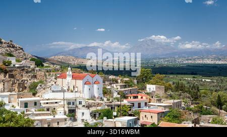 Village traditionnel de Plora, en Crète avec une église blanche et un toit rouge sous un ciel bleu vif, entouré d'oliveraies et de paysages montagneux Banque D'Images