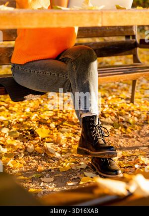 Personne féminine assise à la table dans le parc d'automne portant une tenue saisonnière décontractée. Banque D'Images