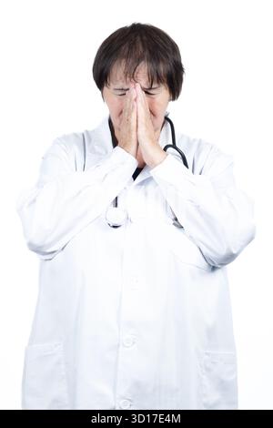 Médecin femme portant une blouse de laboratoire blanche et un stéthoscope éprouvant le stress et l'épuisement professionnel, couvrant son visage avec des mains serrées dans un moment de désespoir Banque D'Images