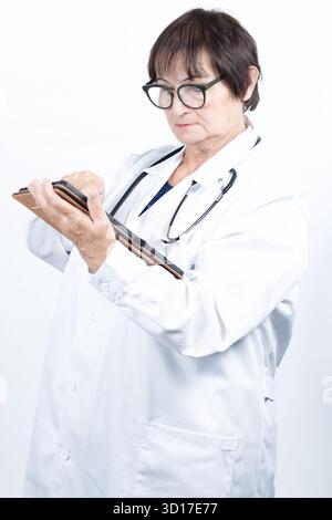 Médecin senior femme portant des lunettes et une blouse de laboratoire blanche, tenant et faisant défiler sur une tablette numérique, gérant les informations du patient Banque D'Images