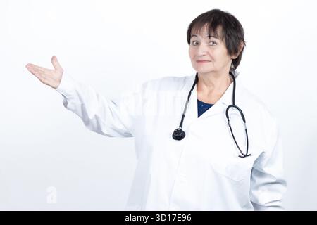 Médecin senior femme portant une blouse de laboratoire et un stéthoscope, souriant tout en faisant des gestes avec une paume ouverte, présentant ou expliquant des concepts Banque D'Images