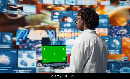 Ordinateur portable à écran vert utilisé par un expert de la santé examinant les tomodensitométries, les données IRM et les analyses de laboratoire sur un mur vidéo. Scientifique dans le laboratoire de recherche médicale lisant des données de santé avec un carnet maquette, caméra A. Banque D'Images