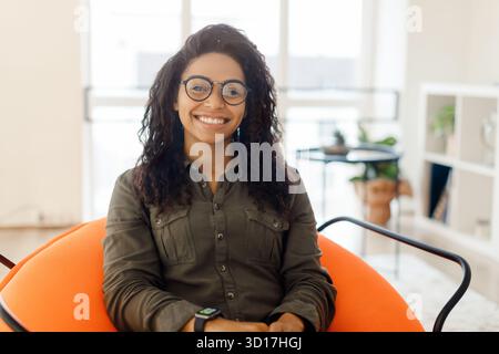 Jeune femme avec cheveux bouclés et lunettes souriant dans une salle moderne et bien éclairée pendant la journée Banque D'Images