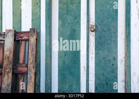 peinte avec des bandes verticales vertes et blanches, la texture montre de la peinture écaillée et est partiellement obstruée par une pile de palettes en bois foncé Banque D'Images