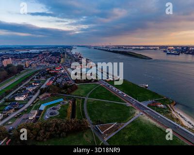Vue aérienne du port fluvial au coucher du soleil des navires de fret, des grues et des quais industriels bordent les deux rives, avec une grille urbaine dense et des champs verts encadrant l'ouate Banque D'Images