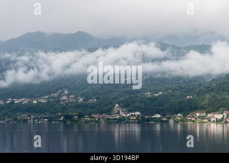 Tôt le matin sur le lac Orta dans le nord de l'Italie, les nuages bas dérivant au-dessus des collines verdoyantes brumeuses et des villages tranquilles reflétés dans la surface calme miroir de wa Banque D'Images