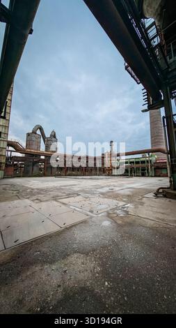 Complexe industriel abandonné avec tuyaux rouillés et cour de béton sous ciel nuageux. Banque D'Images