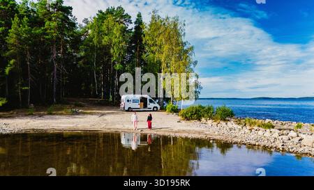 Un couple d'hommes et de femmes se tiennent sur la rive tranquille du lac Saimaa Finlande, entouré d'arbres luxuriants et d'une vue panoramique. Un camping-car est garé à proximité, invitant à l'aventure et à l'exploration. Banque D'Images