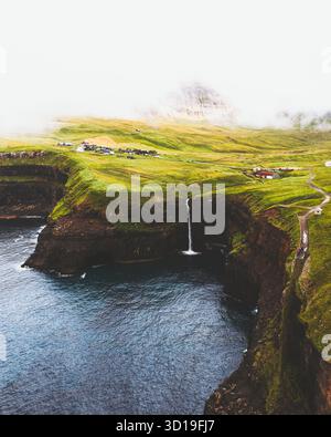 Vue aérienne de la cascade de Múlafossur plongeant dans l'océan sombre depuis un village verdoyant à flanc de falaise entouré de brume, cascade de Múlafossur, Vágar, îles Féroé. Banque D'Images