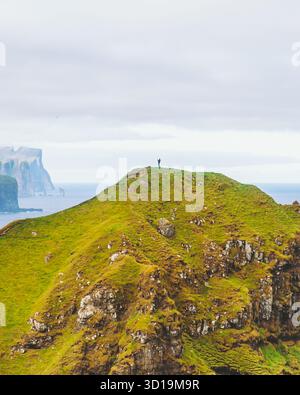 Vue d'une figure solitaire se dresse au sommet d'un pic vert vibrant, surplombant le vaste océan et les falaises spectaculaires sous un ciel nuageux, phare de Kallur, Norð Banque D'Images