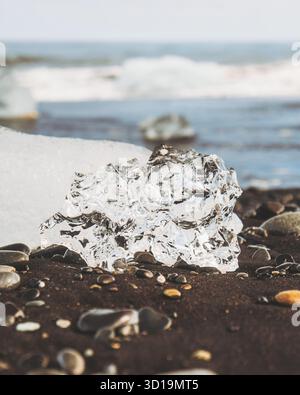 Vue d'un morceau de glace scintillant reposant sur la plage de sable noir contrastant avec les vagues bleues lointaines, Diamond Beach, Sveitarfélagið Hornafjörður, Islande. Banque D'Images
