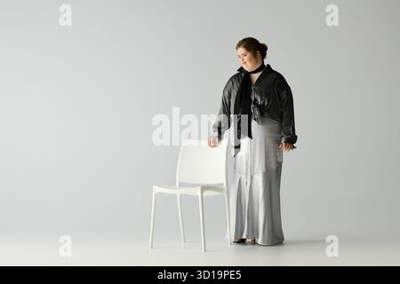 La jeune femme exprime son individualité avec grâce tout en se tenant debout à côté d'une chaise blanche. Banque D'Images