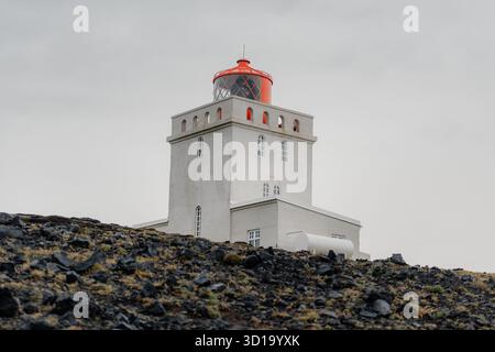 Vue d'un phare blanc avec un haut rouge vif debout contre un ciel silencieux, perché au sommet d'une colline rocheuse accidentée et sombre, plage de Reynisfjara, Mýrdalshreppur, Islande. Banque D'Images