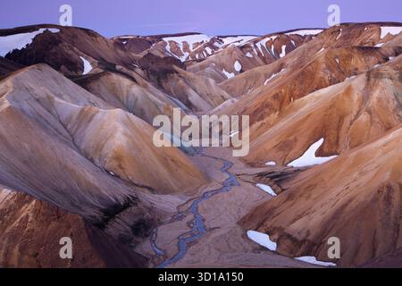 Vue d'une rivière glaciaire sinueuse coupant à travers les montagnes de rhyolite aux couleurs multiples sous un ciel violet, coiffé de neige, Landmannalaugar, Rangárþing ytra, Islande. Banque D'Images