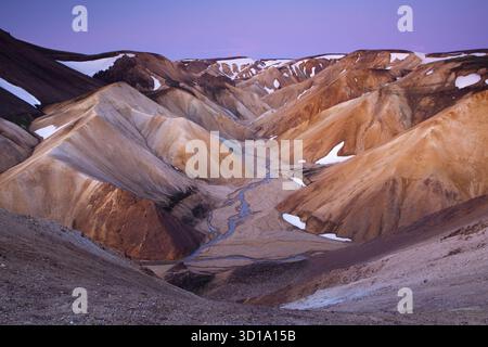 Vue sur les montagnes colorées des Highlands islandais et les zones glacées, avec une rivière sinueuse coupant la vallée sous un ciel crépusculaire, Landmannalaugar, Rangárþing ytra, Islande. Banque D'Images
