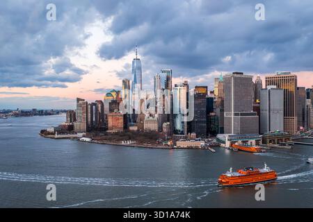 Vue aérienne des gratte-ciel FIDI perce le ciel près du One World Trade Center, avec un ferry coupant à travers les eaux sombres sous une aube pastel, New York, New York, États-Unis. Banque D'Images