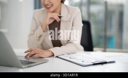 Souriante femme d'affaires adulte asiatique effectue des tâches sur ordinateur portable dans l'espace de travail contemporain. Elle élabore des stratégies pour les transactions immobilières et la sensibilisation des clients. Achie Banque D'Images