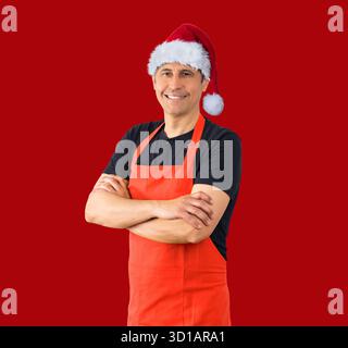 Homme hispanique joyeux homme positif souriant dentiellement avec les bras croisés exprimant, émotions positives tout en portant un chapeau de Noël contre un dos rouge Banque D'Images